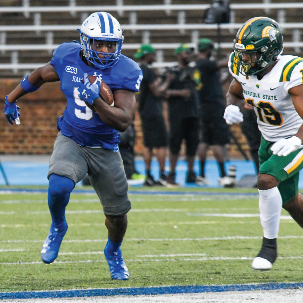 Hampton University football player running with the ball during a game while pursued by a Norfolk State defender on the field.