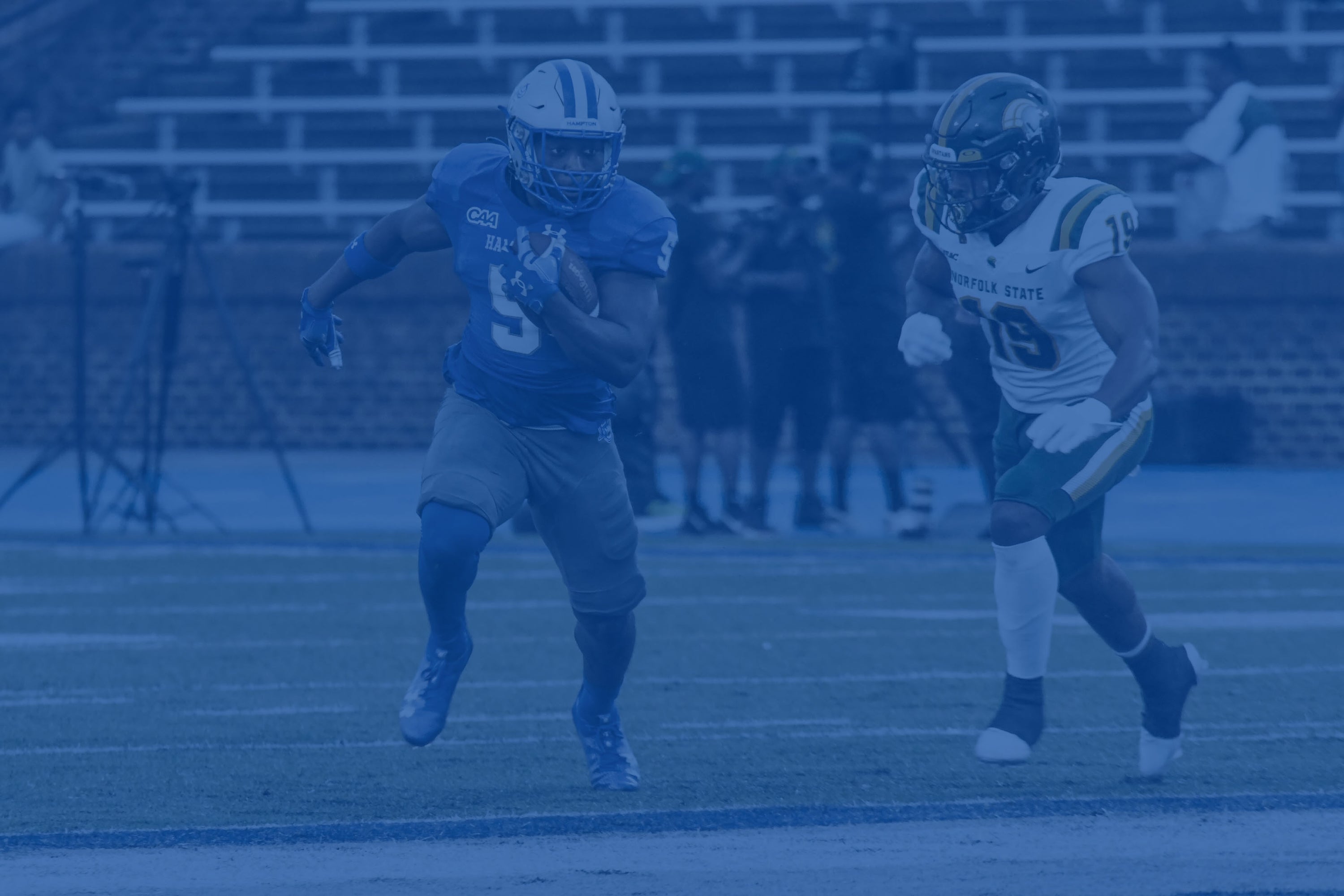 Action shot of two football players competing on the field. A Hampton University player carries the football. The image is covered with a blue color overlay.