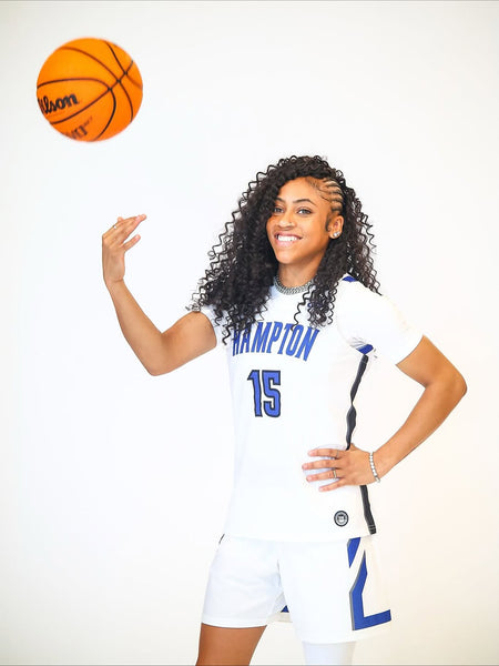 Hampton University women’s basketball player in white uniform smiling while spinning a basketball against a light background.