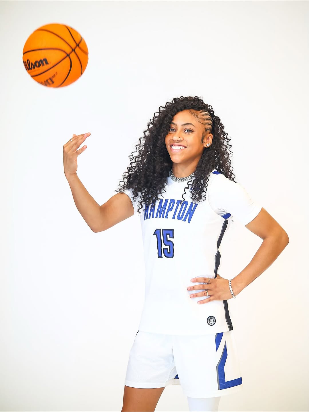 Hampton University women’s basketball player in white uniform smiling while spinning a basketball against a light background.