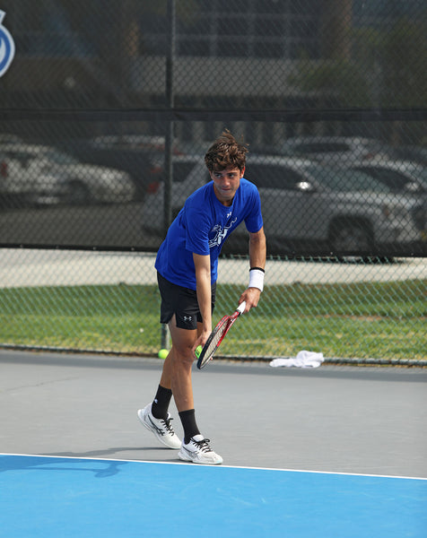 Hampton U male tennis player about to serve on a court with cars in the background
