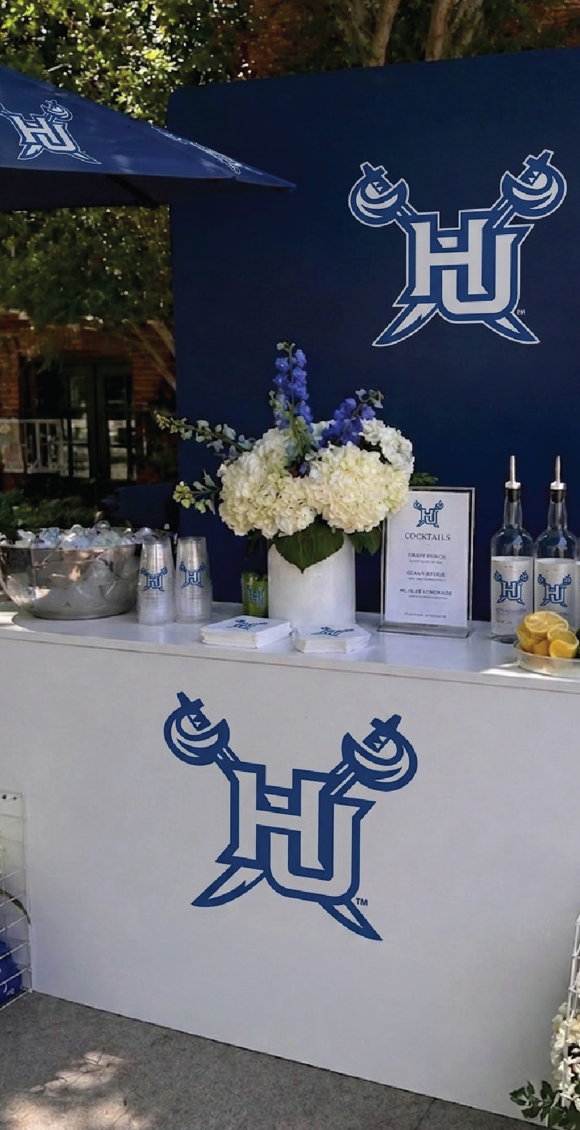 A branded outdoor beverage station with a white counter and dark blue backdrop, featuring the HU logo, floral centerpieces, bottles, ice bucket, and signage.