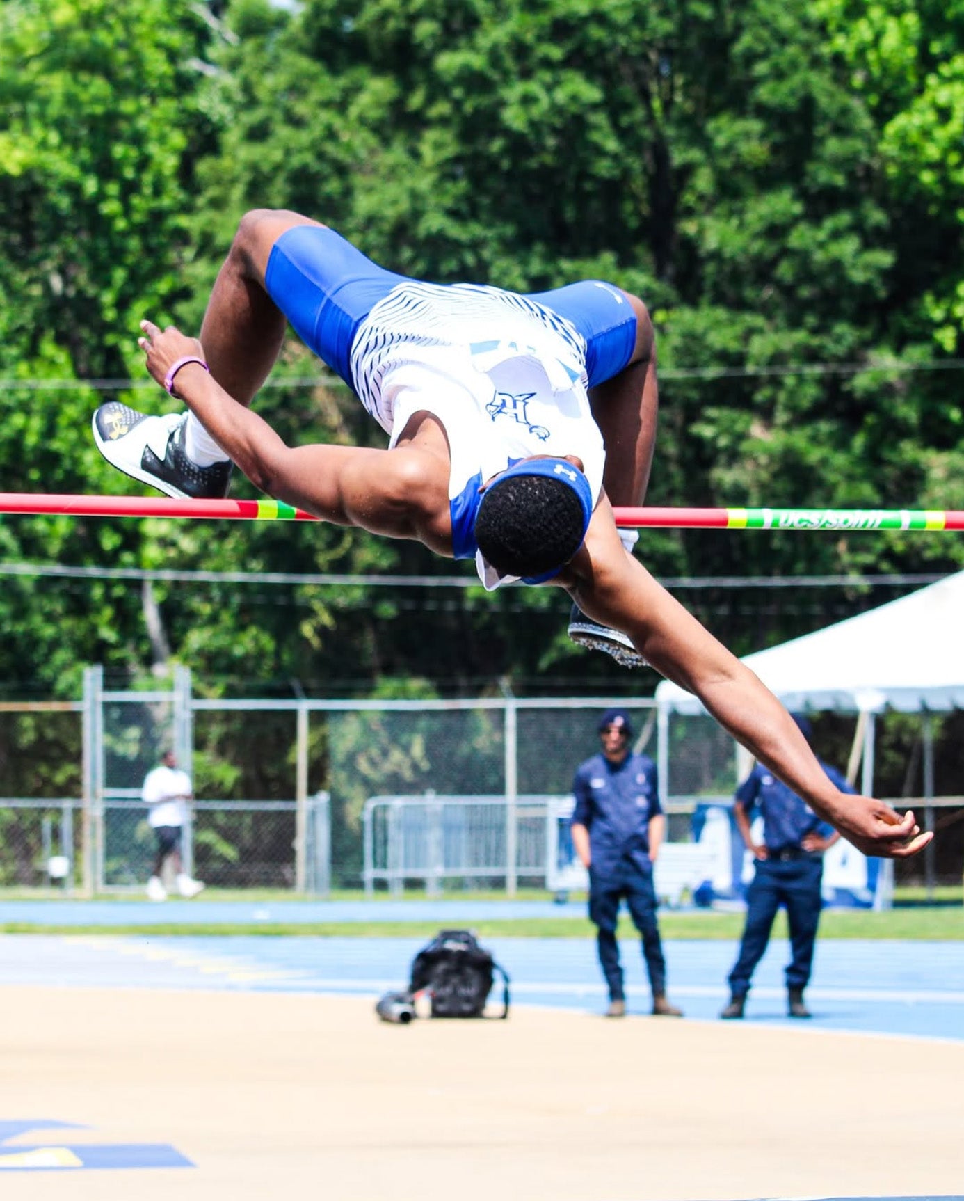 Athlete performing a high jump on a track and field event with trees in the background.