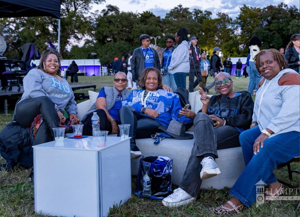 A group of five people sitting together on a white outdoor sofa at an event, wearing matching blue and white graphic sweatshirts. A small white side table with drinks is in the foreground, with trees and other event attendees in the background.