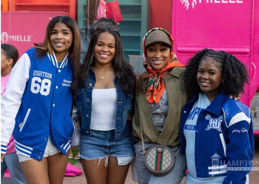 Four women standing together in front of a bright pink truck or booth, wearing varsity jackets and casual outfits. Visible text includes 'MIEILLE' on the pink vehicle and 'HAMPTON' on a jacket.
