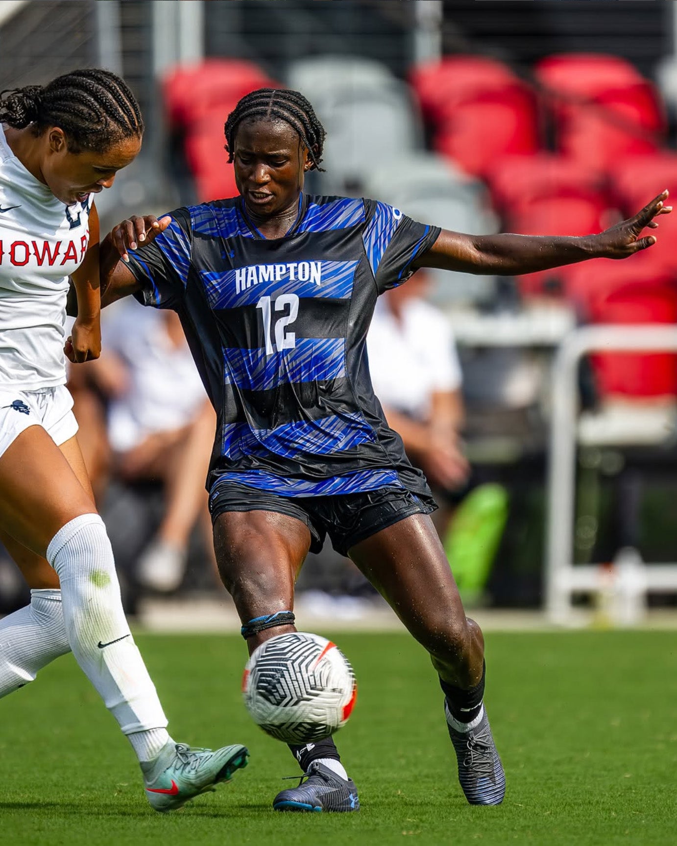 Two female soccer players in action on a field with stadium seats in the background.